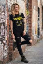 Woman leaning against a brick wall outdoors wearing a black cotton Nirvana graphic t-shirt with a vintage distressed smiley face