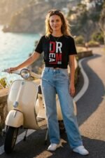 Woman leaning against a vintage scooter outdoors wearing a black cotton t-shirt with a vertical Dream Big typography graphic