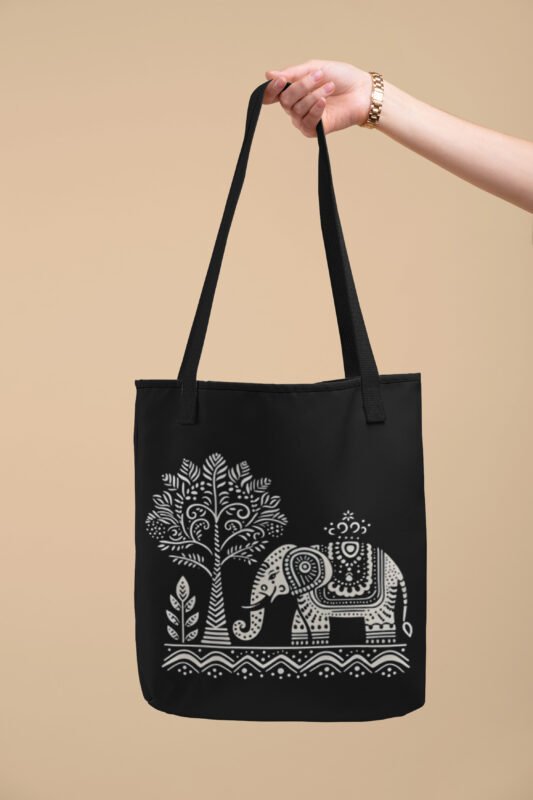 Black canvas tote bag with a white tribal-style elephant and tree design, held by a hand against a beige background.