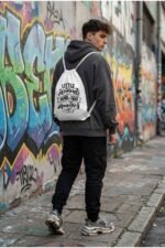 Back view of a model wearing Stilotee white canvas drawstring backpack with minimalist typography print against a colorful urban graffiti wall