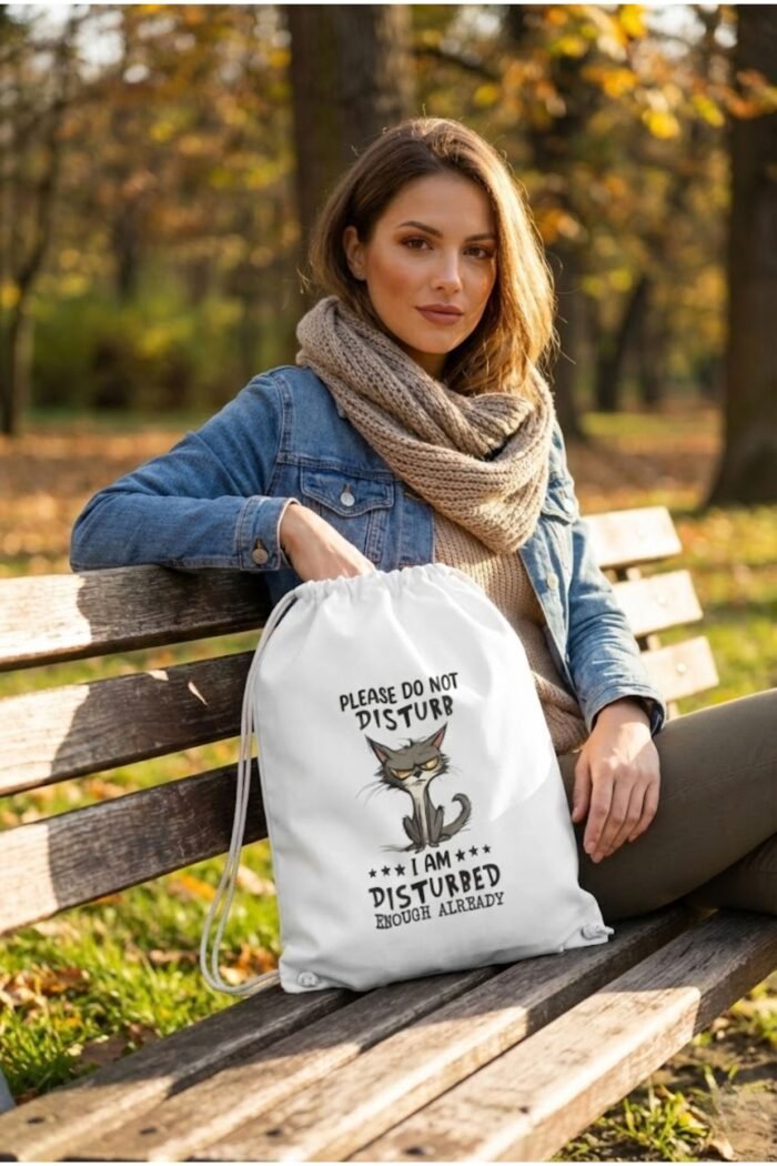 A female model sitting on a concrete ledge outdoors, wearing the white sarcastic cat drawstring sackpack on her back.