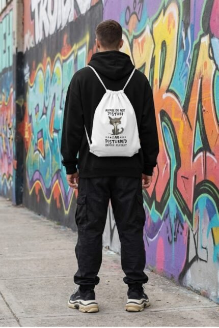 A streetwear mockup of a person in a black hoodie wearing the white canvas drawstring backpack with the angry grey cat graphic.