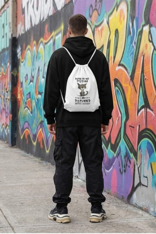 A streetwear mockup of a person in a black hoodie wearing the white canvas drawstring backpack with the angry grey cat graphic.