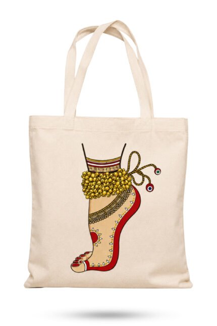 Off-white canvas tote bag featuring an Indian classical dancer foot adorned with golden ghungroo bells, red alta dye, and ornamental anklet with zipper closure.