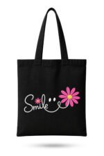 Black heavyweight canvas tote bag featuring white cursive Smile text with pink daisy flowers and smiley face graphic with zipper closure.